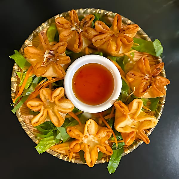 Golden fried Thai crab rangoons arranged on a woven plate with lettuce, carrots, and dipping sauce.