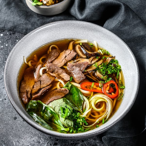 A bowl of Thai beef noodle soup with mushrooms, green onions, and red chili.